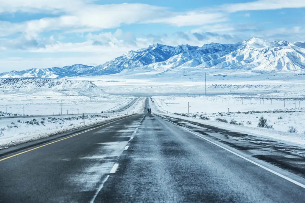 A long, straight highway stretches into the distance through a snow-covered landscape, leading toward distant mountains under a cloudy sky.