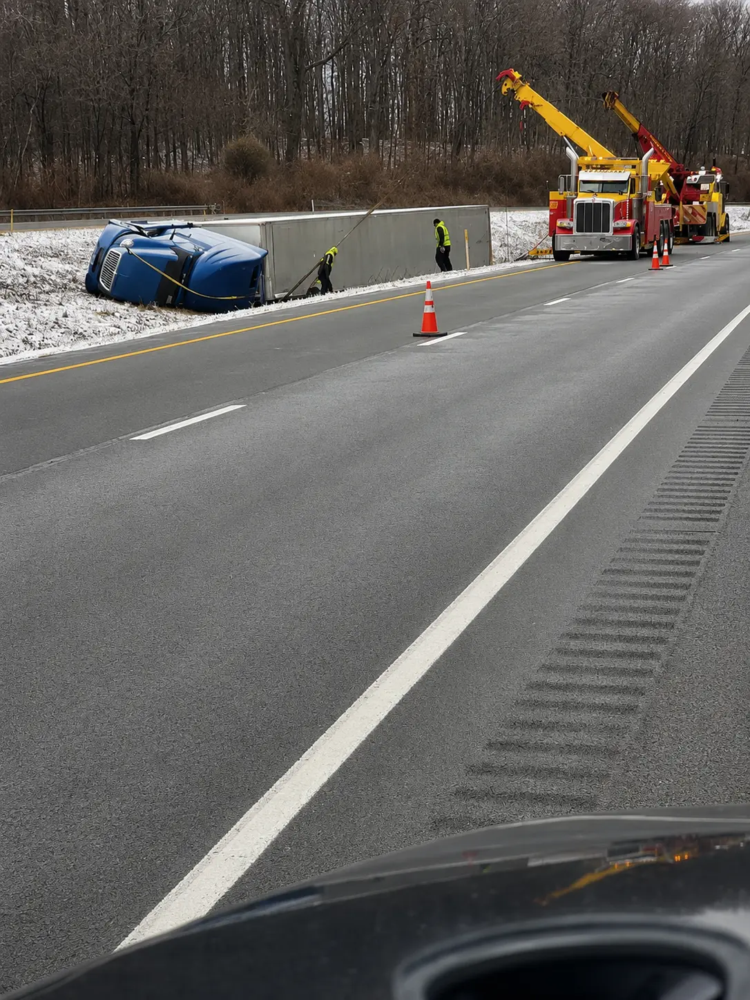 A blue car is overturned in a snowy ditch beside a highway. Two tow trucks and workers with safety vests are on the scene, preparing to recover the vehicle. Traffic cones line the roadside. Trees are visible in the background.