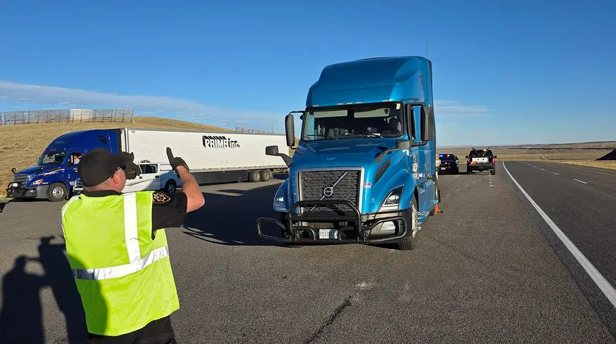 A person in a yellow safety vest directs a blue semi-truck on the side of a highway. Other trucks and vehicles are parked nearby on a clear day with a blue sky.