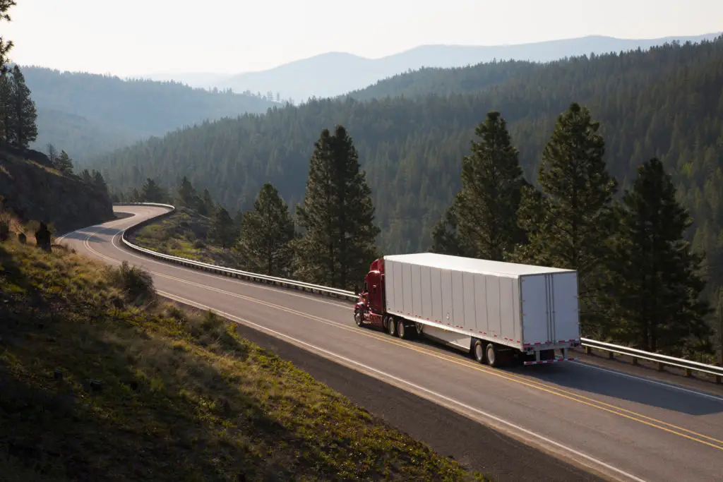 A red semi-truck with a white trailer drives along a winding mountain road surrounded by pine trees and rolling hills under a hazy sky.