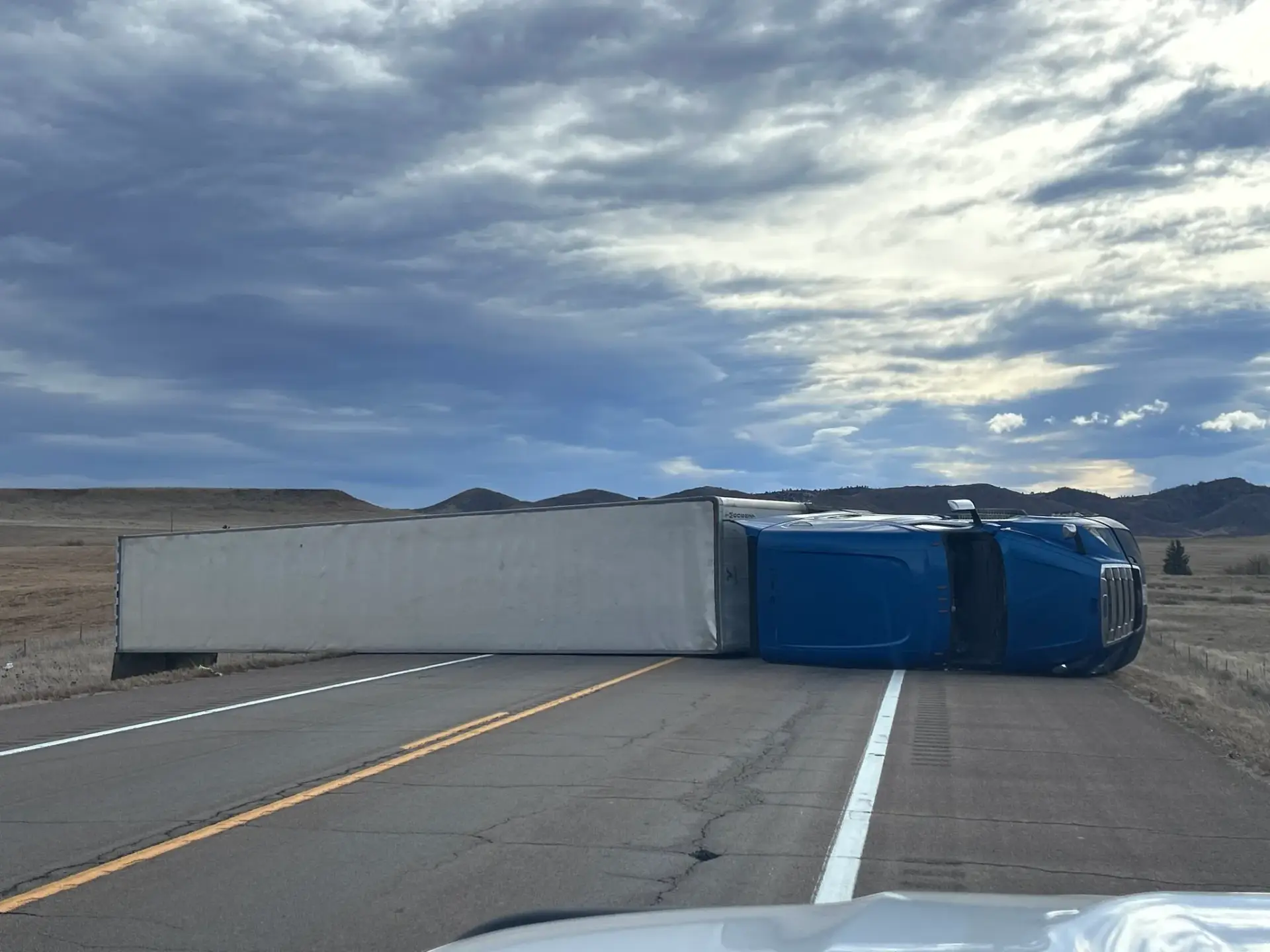 A blue semi-truck with a white trailer is overturned, blocking both lanes of an empty rural road under a cloudy sky, with open fields and distant hills in the background.