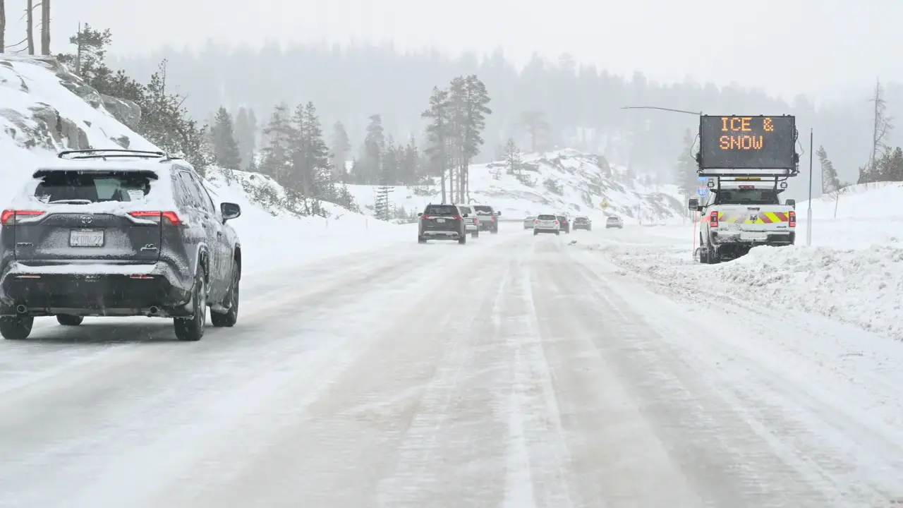 Cars drive on a snow-covered road surrounded by trees, with a digital road sign on the right displaying a warning about ICE & SNOW. The scene is overcast and visibility appears reduced due to wintry conditions.
